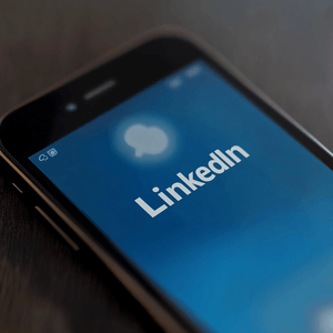 Anunciar-no-linkedin Celular exibindo tela do LinkedIn com destaque para ícone de anúncios e perfil profissional